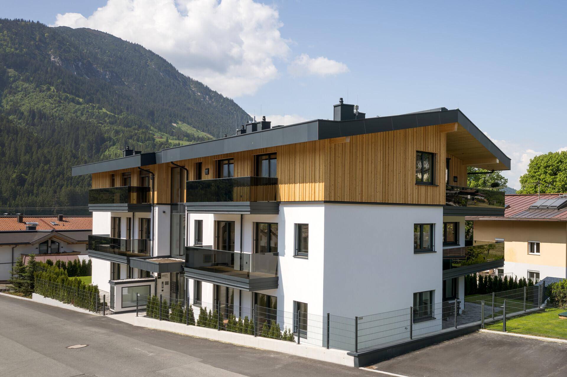 Elegantes Apartmentgebäude mit weißer Fassade, Holzverkleidung und Balkonen vor alpiner Kulisse.