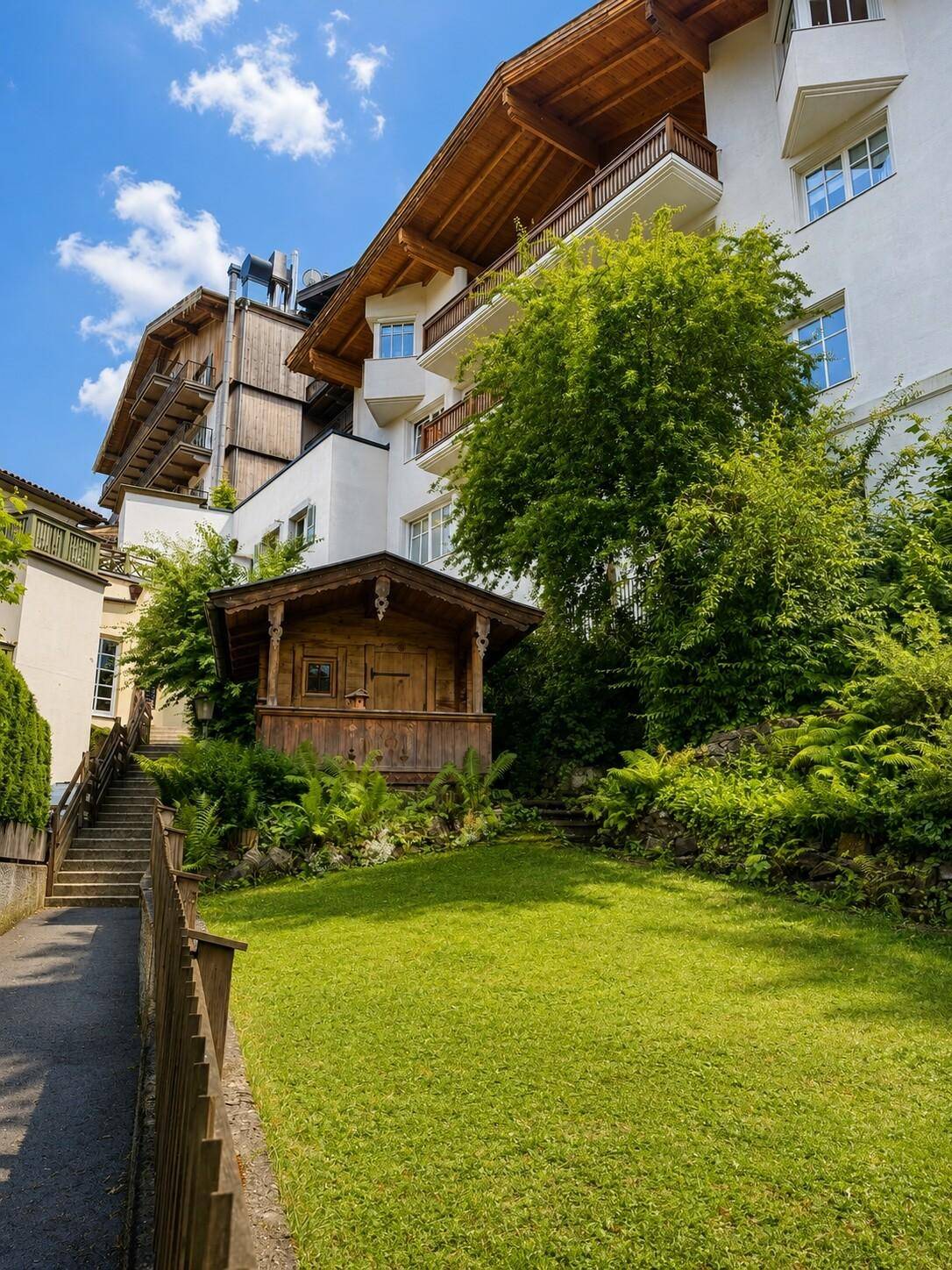 Grüner Gartenbereich mit Rasen und einem charmanten Holzhaus, umgeben von Bäumen und einem größeren Gebäude.