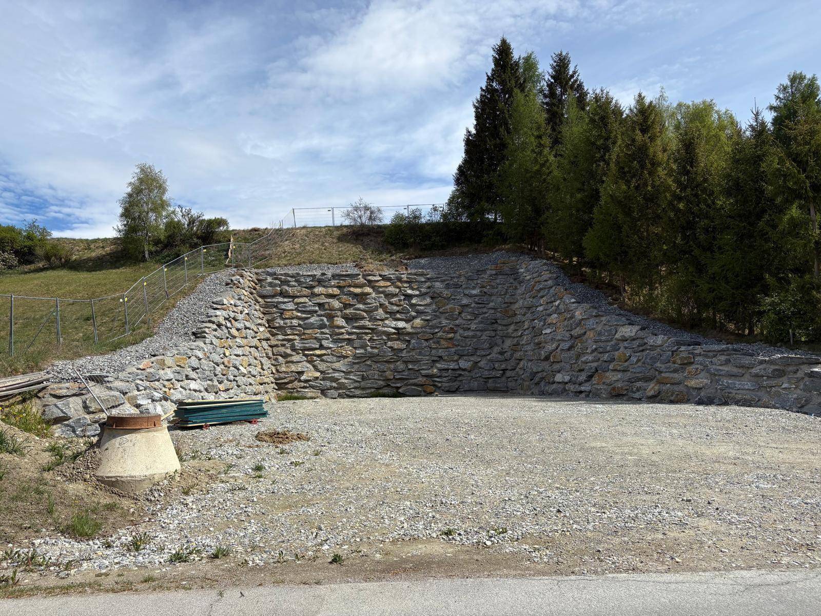 Detailansicht der Natursteinmauer und Schotterfläche mit einem Betonschacht im Vordergrund.