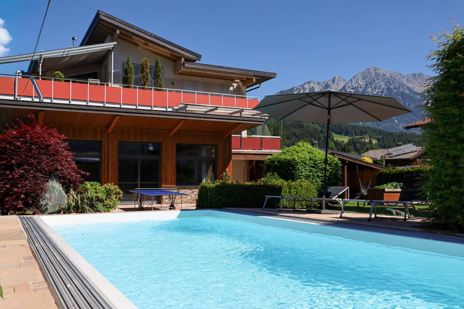 Außenansicht eines modernen Hauses mit großem Pool, Liegestühlen und Sonnenschirm vor Bergkulisse.