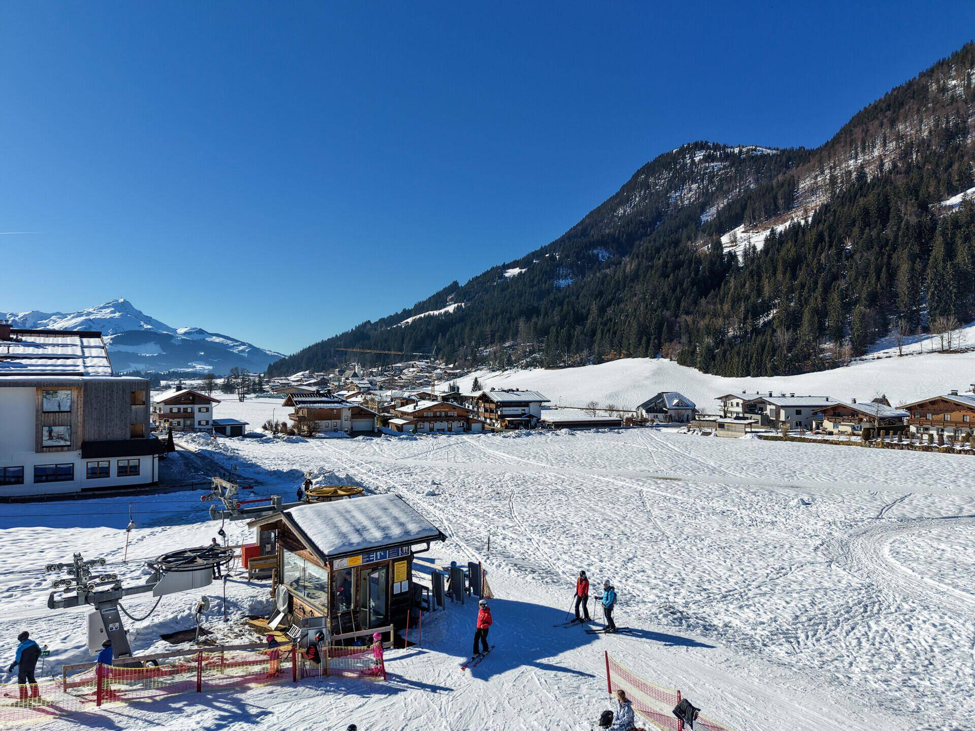 Kleiner Skiliftbereich mit Skifahrern und verschneiter Berglandschaft im Hintergrund an einem sonnigen Tag.