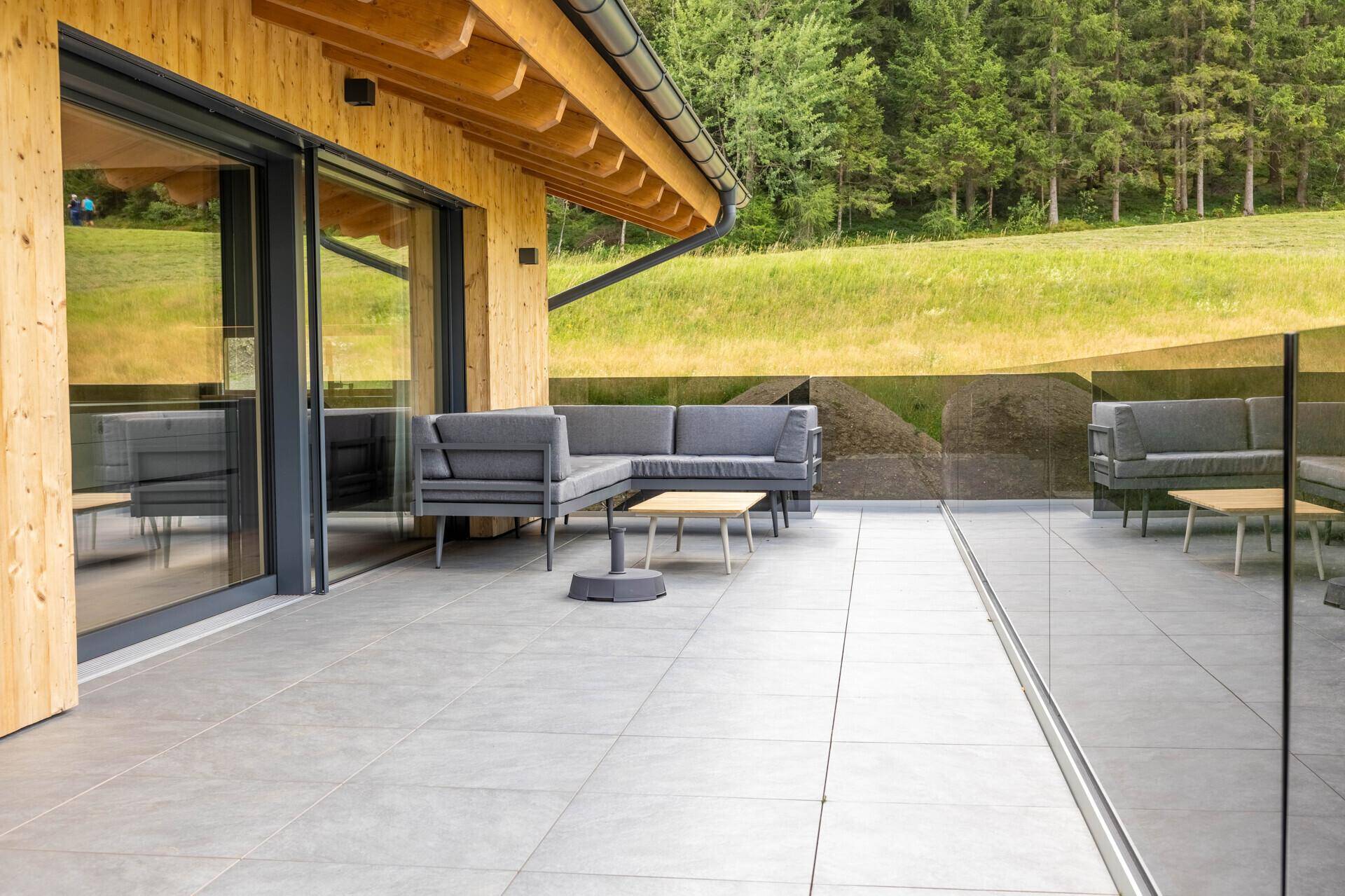 Moderne Terrasse mit bequemen Gartenmöbeln und Holzelementen, umgeben von einem Glasgeländer, bietet einen idyllischen Blick ins Grüne und auf den Wald.