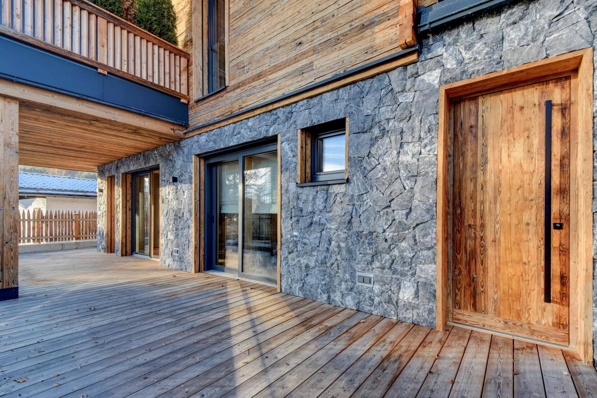 Große Holzterrasse am Erdgeschoss mit Zugang zum Haus und Steinfassade.