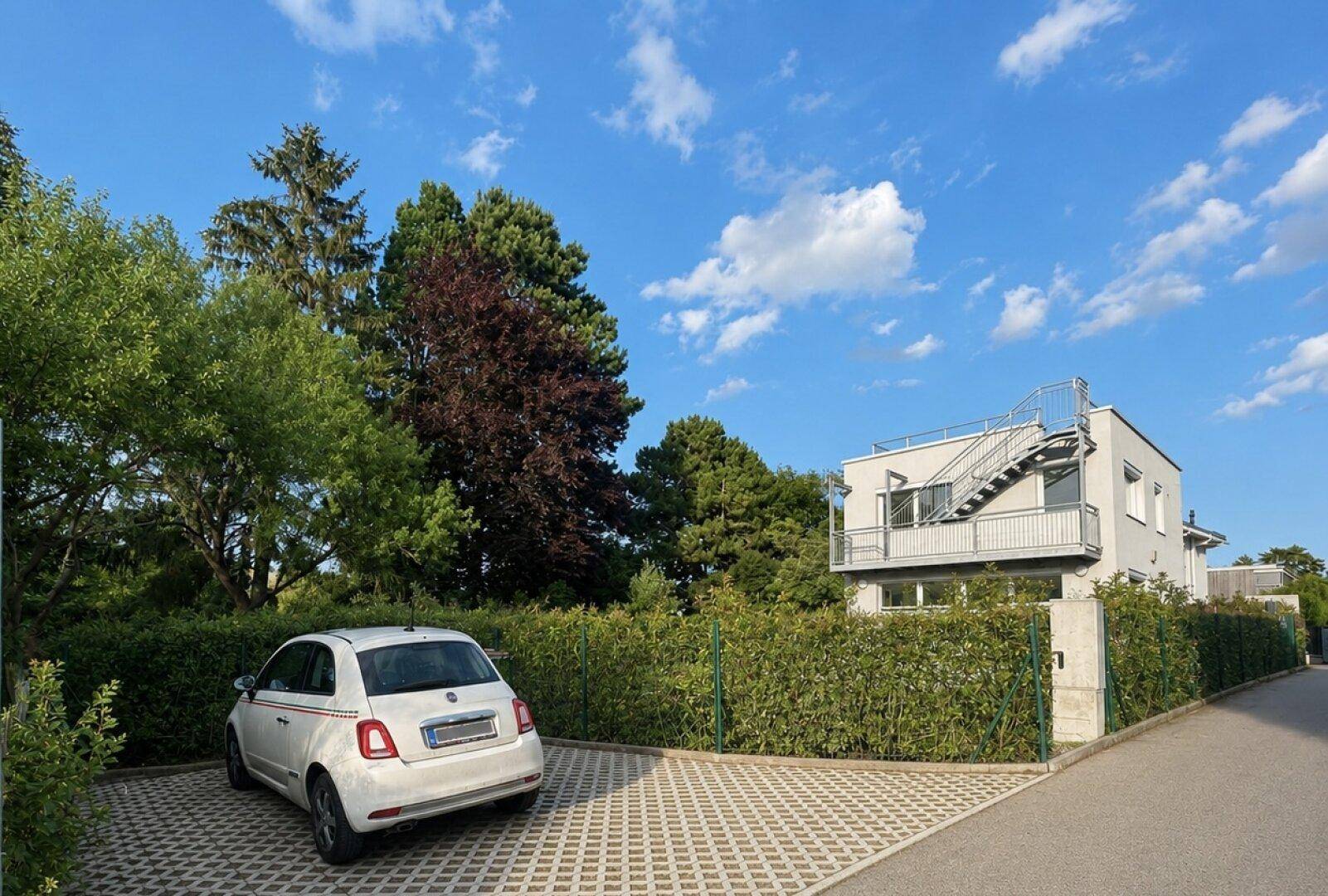 Außenansicht mit einem gepflasterten Parkplatz, umgeben von Hecken und dem modernen Haus im Hintergrund.