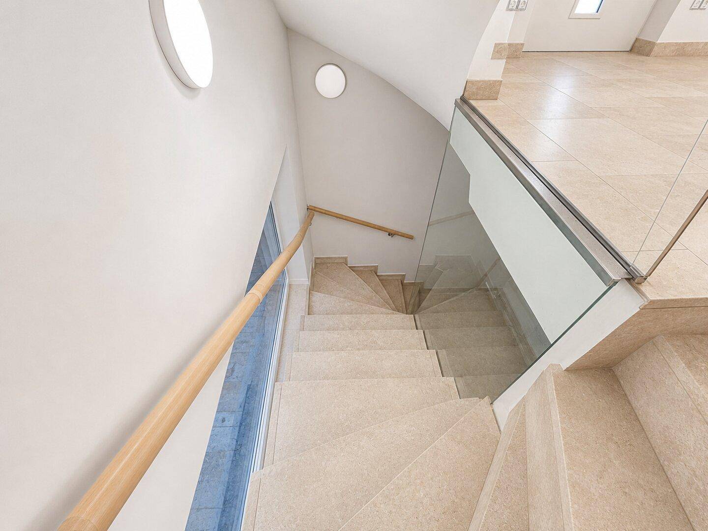 Elegante Treppe mit Fliesenstufen, Glasgeländer und Holzhandlauf, beleuchtet durch runde Fenster.