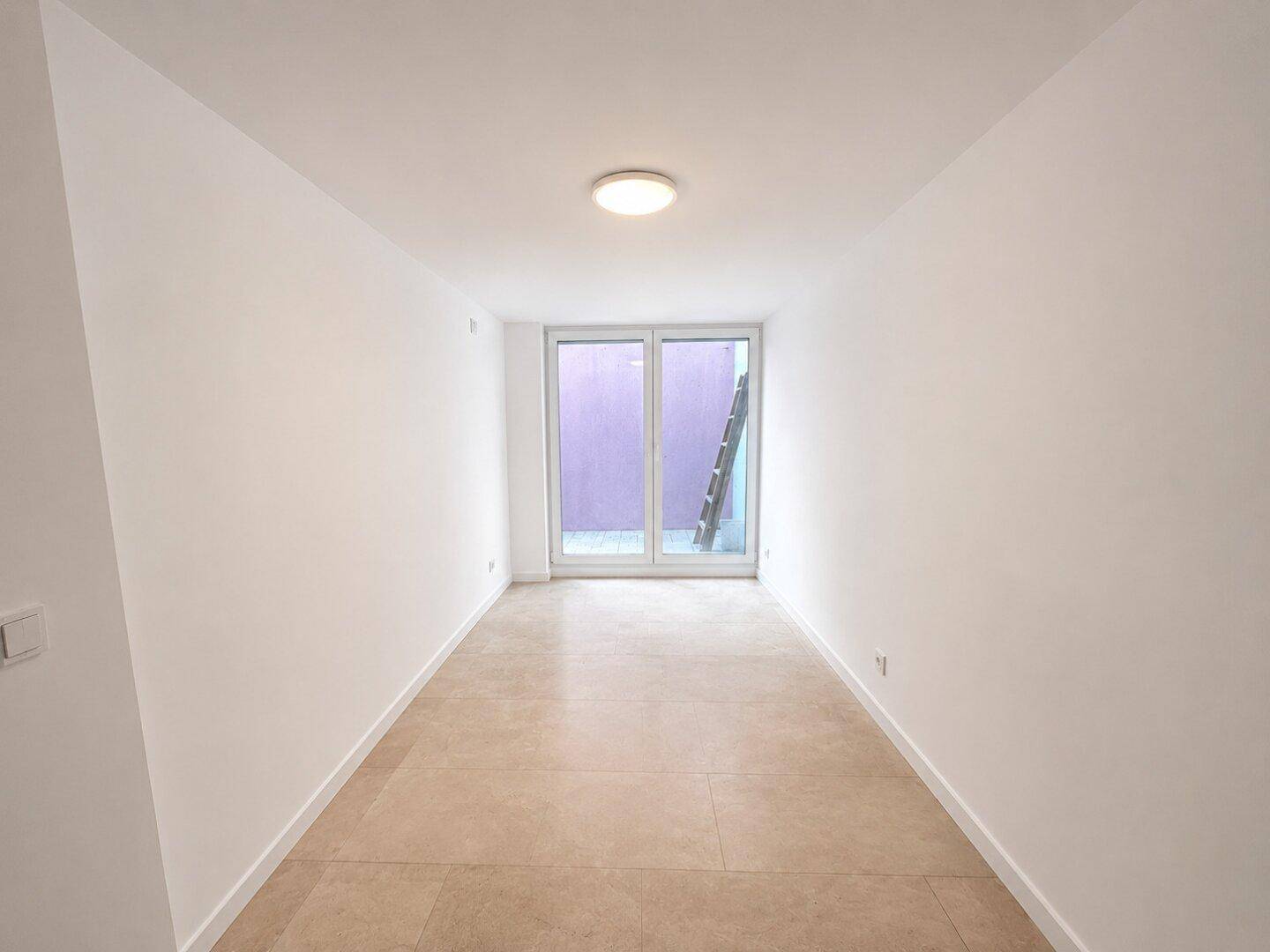 Schmaler Raum mit Fliesenboden und einer Glasschiebetür am Ende, die viel Licht spendet.