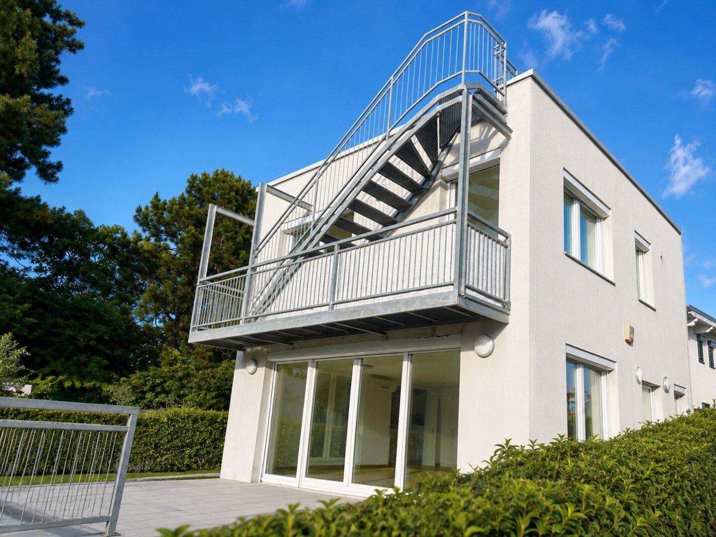 Seitenansicht des modernen Gebäudes mit Außentreppe, Balkon und gepflegter Hecke.
