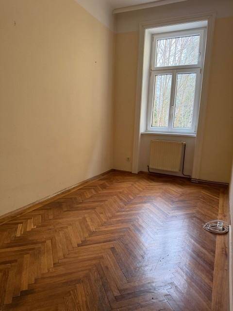Helles Schlafzimmer mit Fischgrätparkett und einem großen Fenster, das viel Tageslicht hereinlässt.