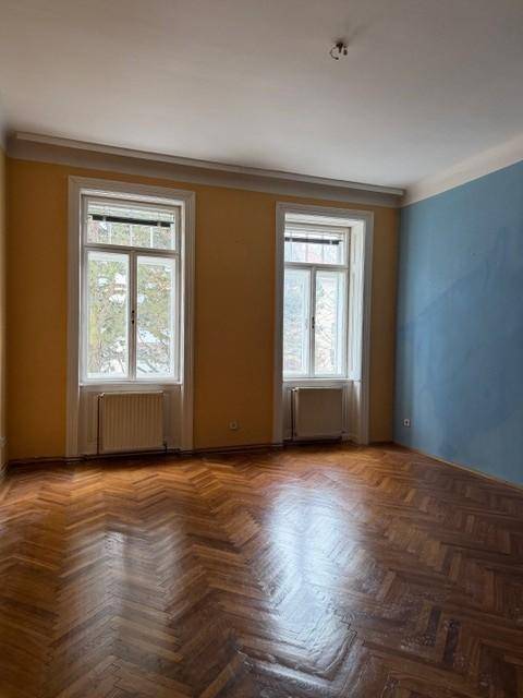 Helles Zimmer mit Fischgrätparkett, zwei hohen Fenstern und Heizkörpern unter den Fenstern.