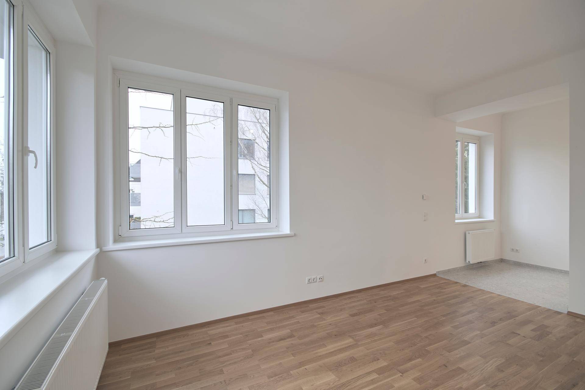 Geräumiger, heller Raum mit Holzparkett und großen Fenstern, der zu einem weiteren Bereich führt.