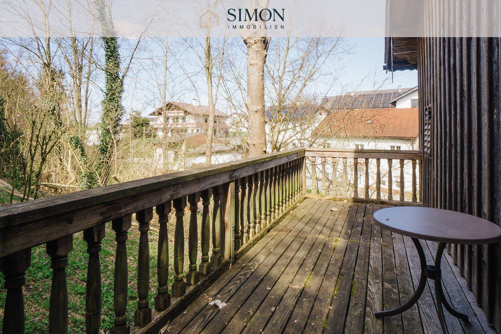 Rustikaler Holzbalkon mit kleinem Tisch und Blick ins Grüne sowie auf umliegende Häuser.