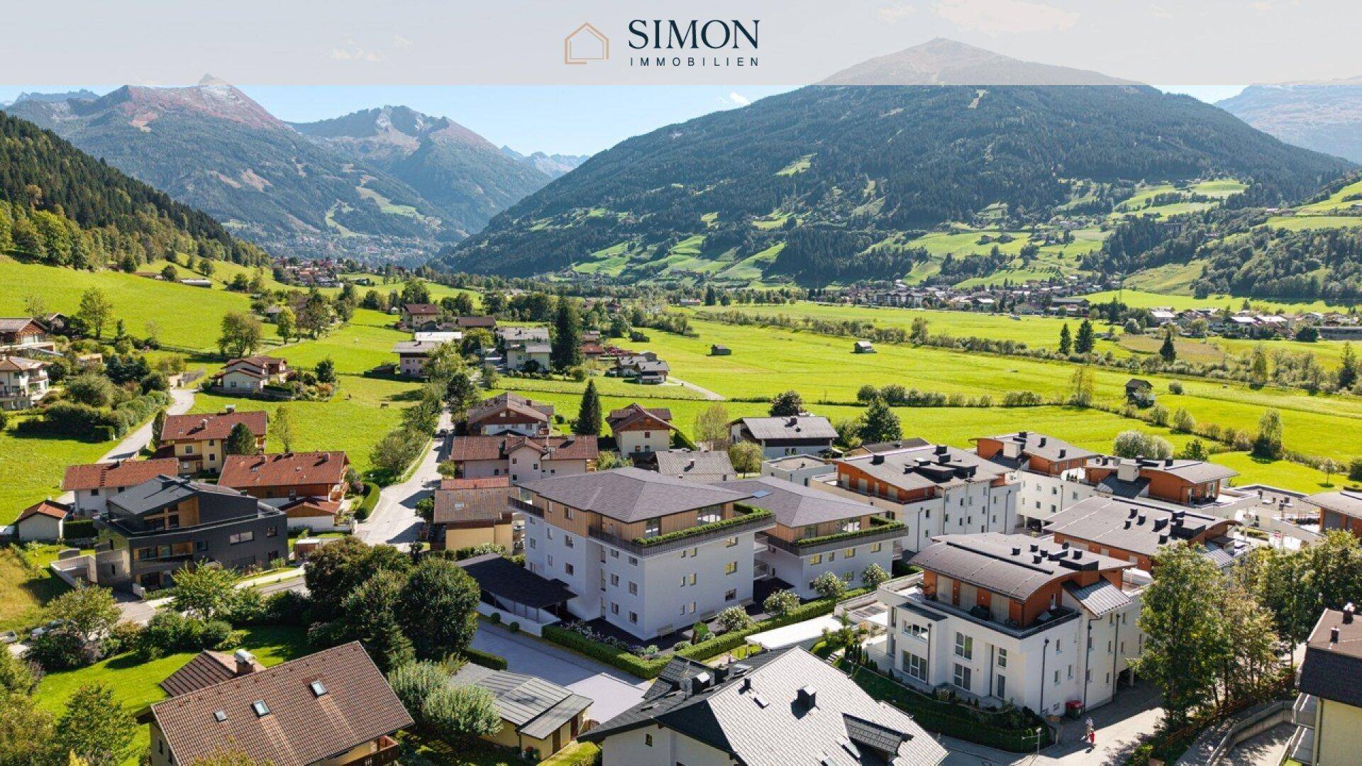 Luftaufnahme der Wohnanlage eingebettet in eine grüne alpine Umgebung mit umliegenden Häusern und Bergen.