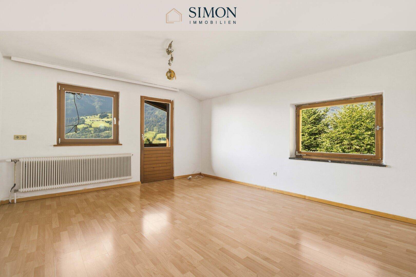 Geräumiges Wohnzimmer mit Holzboden, zwei Fenstern und Zugang zu einem Außenbereich.