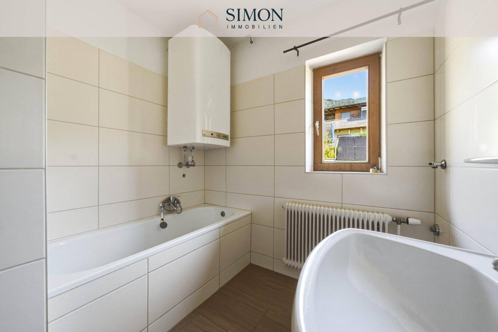 Gefliestes Badezimmer mit Badewanne, Fenster und einem Warmwasserboiler an der Wand.