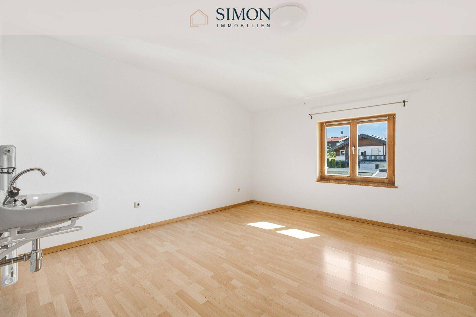 Einfaches Zimmer mit Holzboden, Fenster und einem Waschbecken an der Wand.