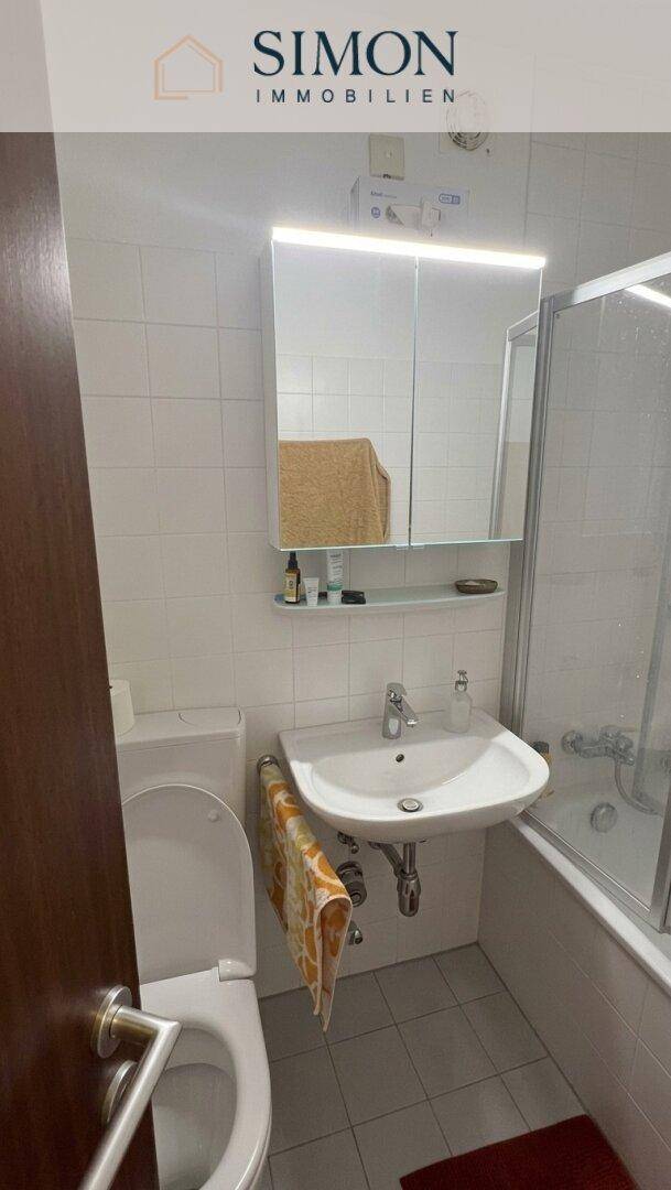 Helles Badezimmer mit weißen Fliesen, Waschbecken, WC und Spiegelschrank über dem Waschtisch.
