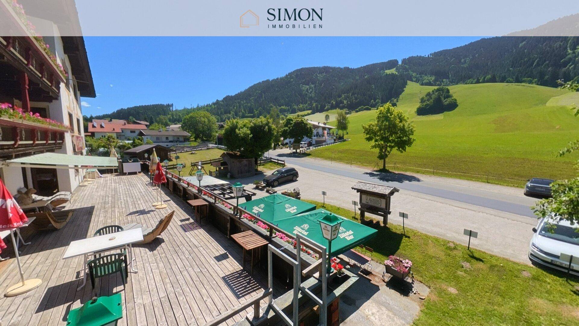 Große Holzterrasse mit Tischen, Stühlen und Sonnenschirmen, bietet einen weiten Blick auf grüne Hügel.