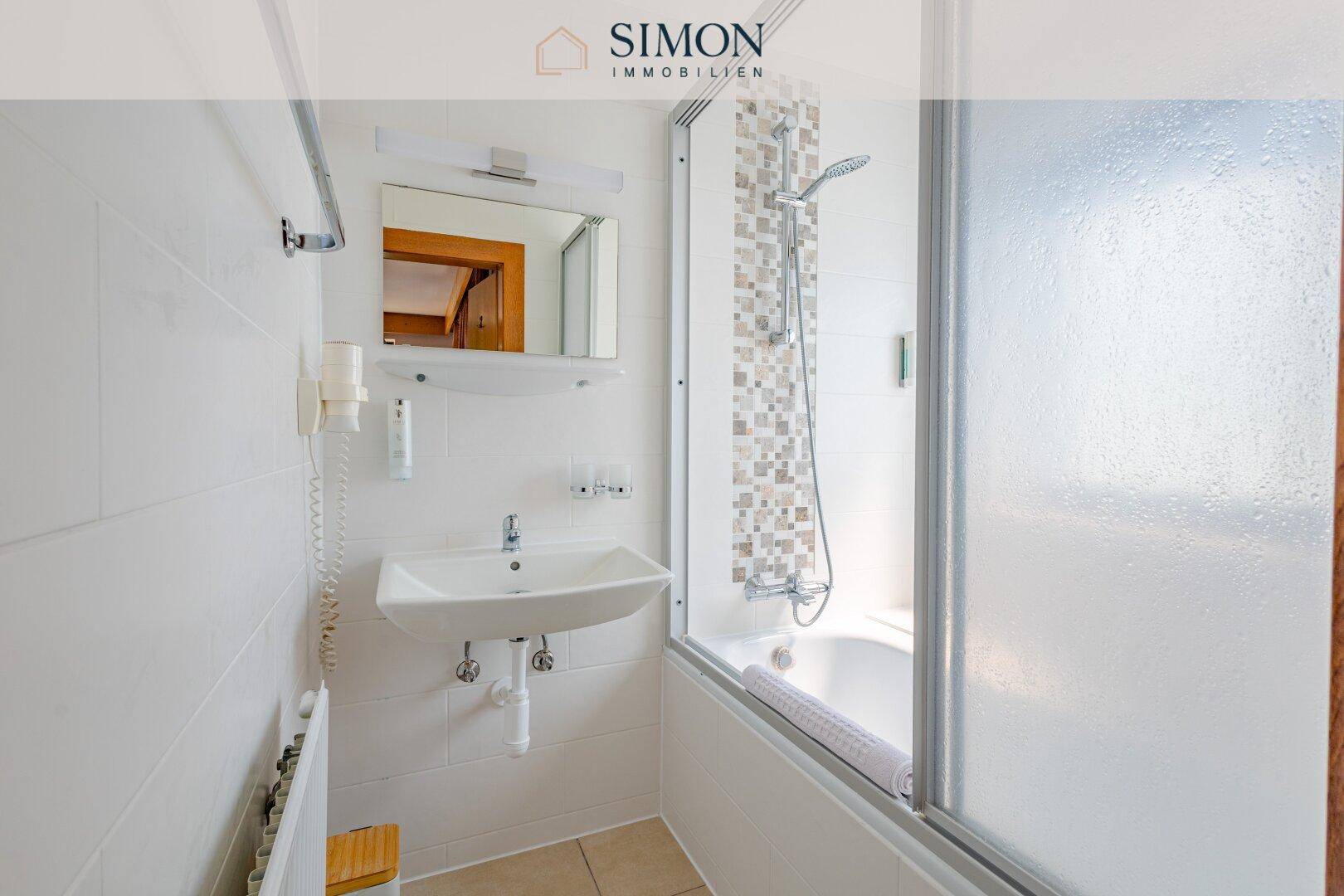 Badezimmer mit Waschbecken, Spiegel und einer Badewanne mit Duschfunktion und Glasabtrennung.