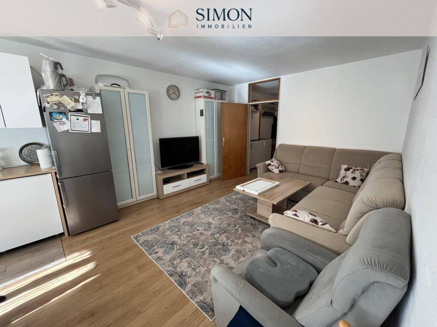 Helles Wohnzimmer mit beigem Ecksofa, Holzboden und integrierter Küchenzeile.