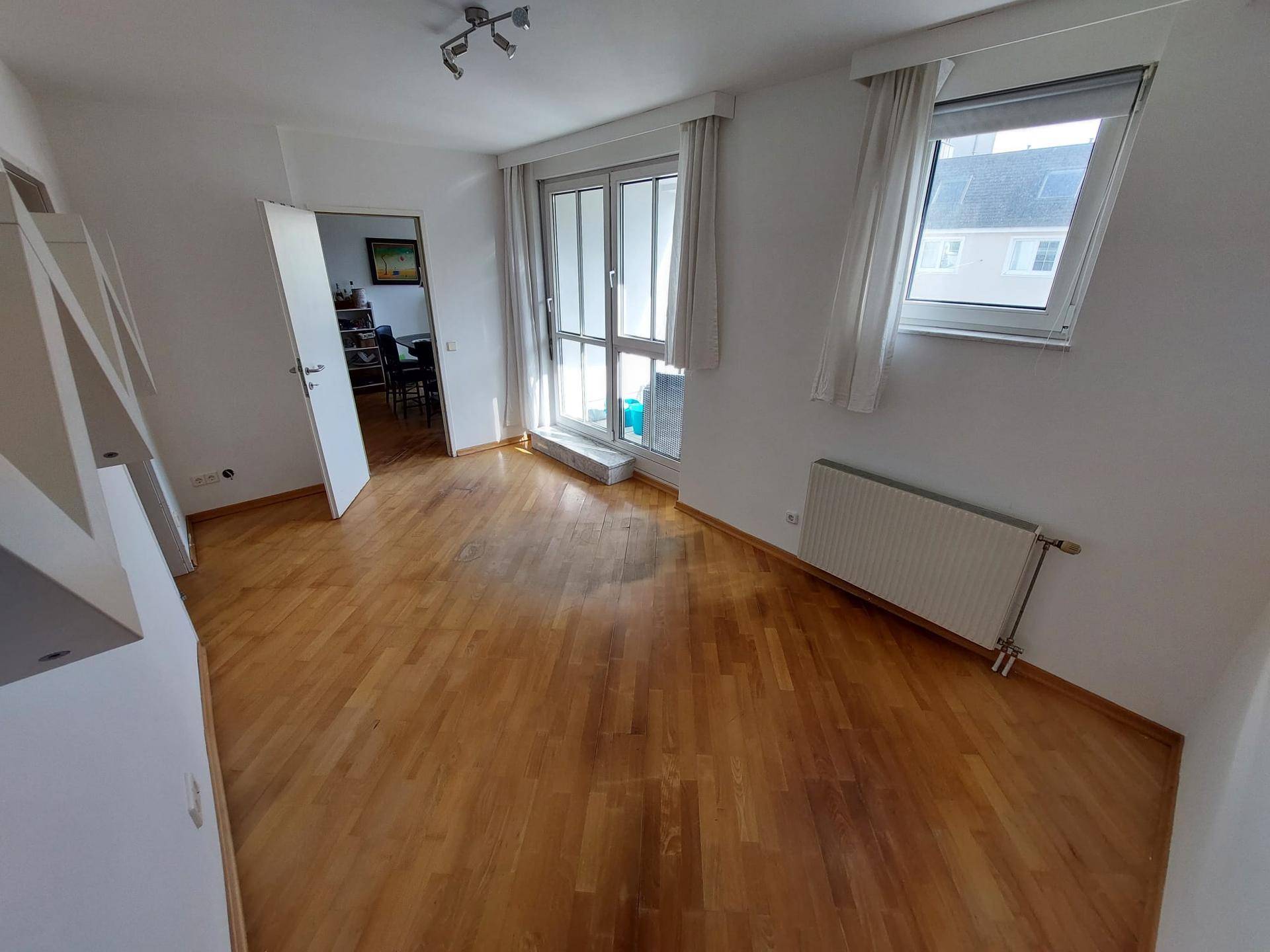 Helles, leeres Zimmer mit Parkettboden, weißen Wänden und Zugang zu einem Balkon.