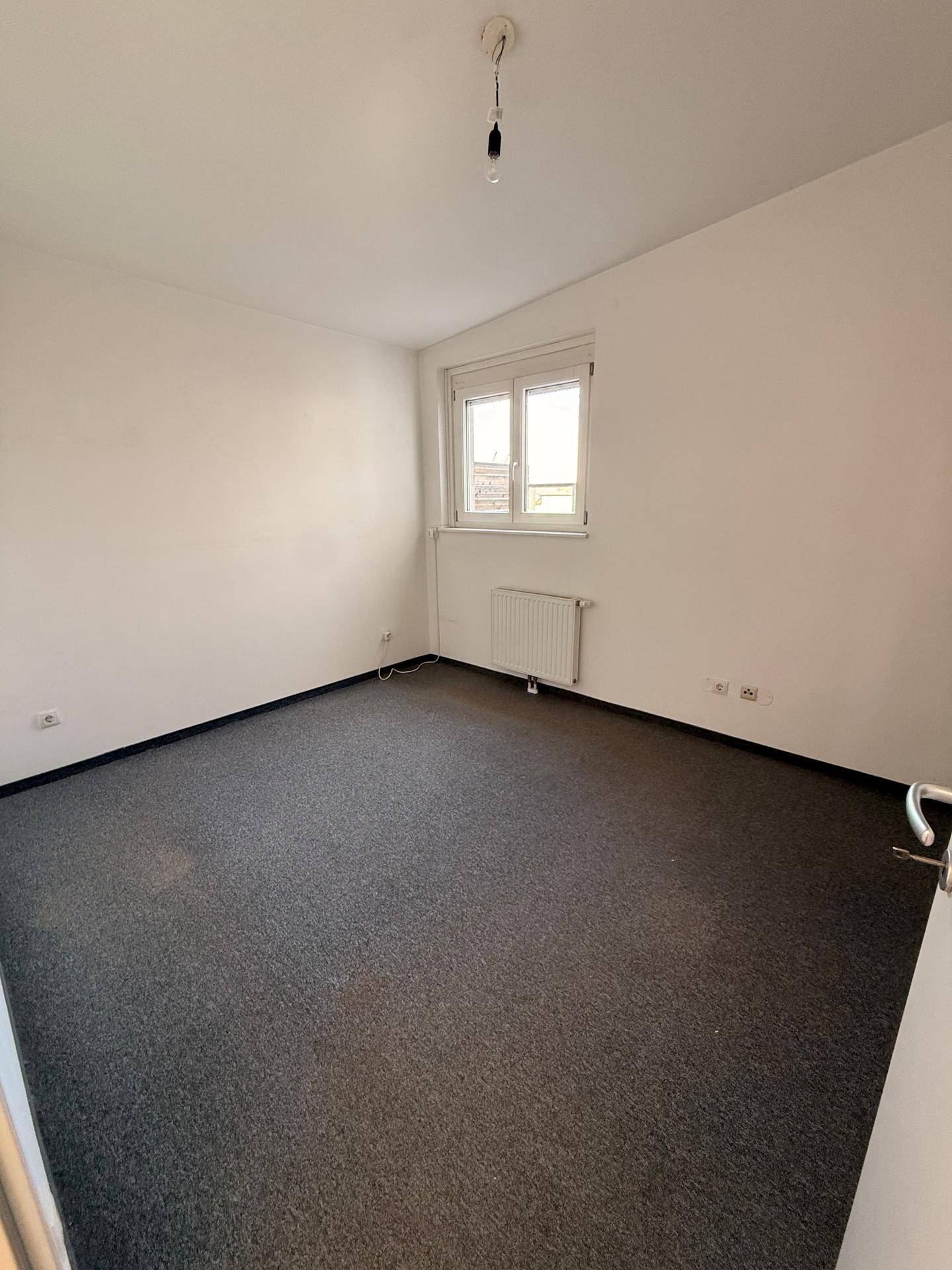 Helles Schlafzimmer mit dunklem Teppichboden und einem Fenster, das den Raum erhellt.