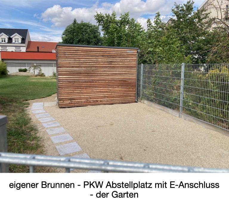 Gartenbereich mit Kiesweg, Holzschuppen, Zaun und einem PKW-Stellplatz mit E-Anschluss.