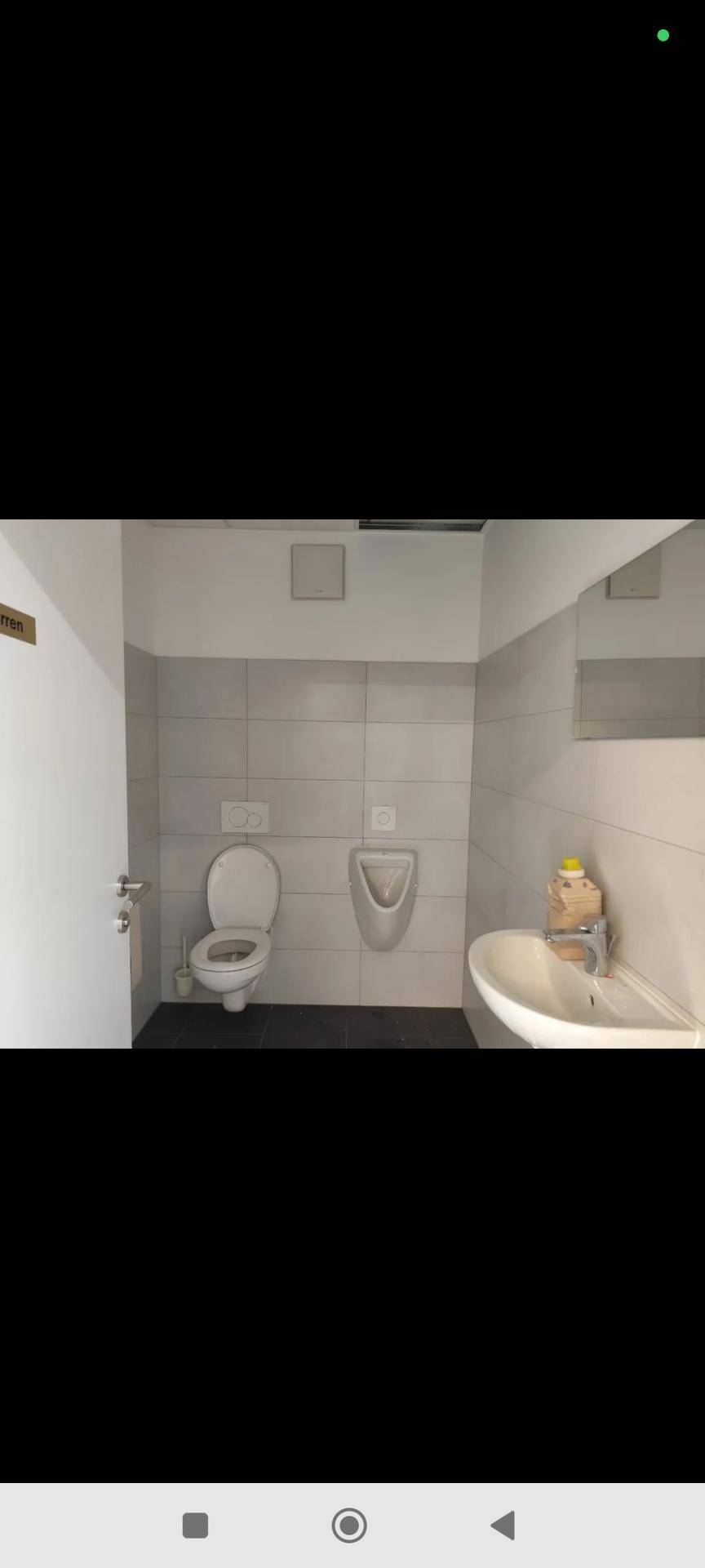 Funktionales Badezimmer mit hellgrauen Fliesen, einer Toilette, einem Urinal und einem Waschbecken.