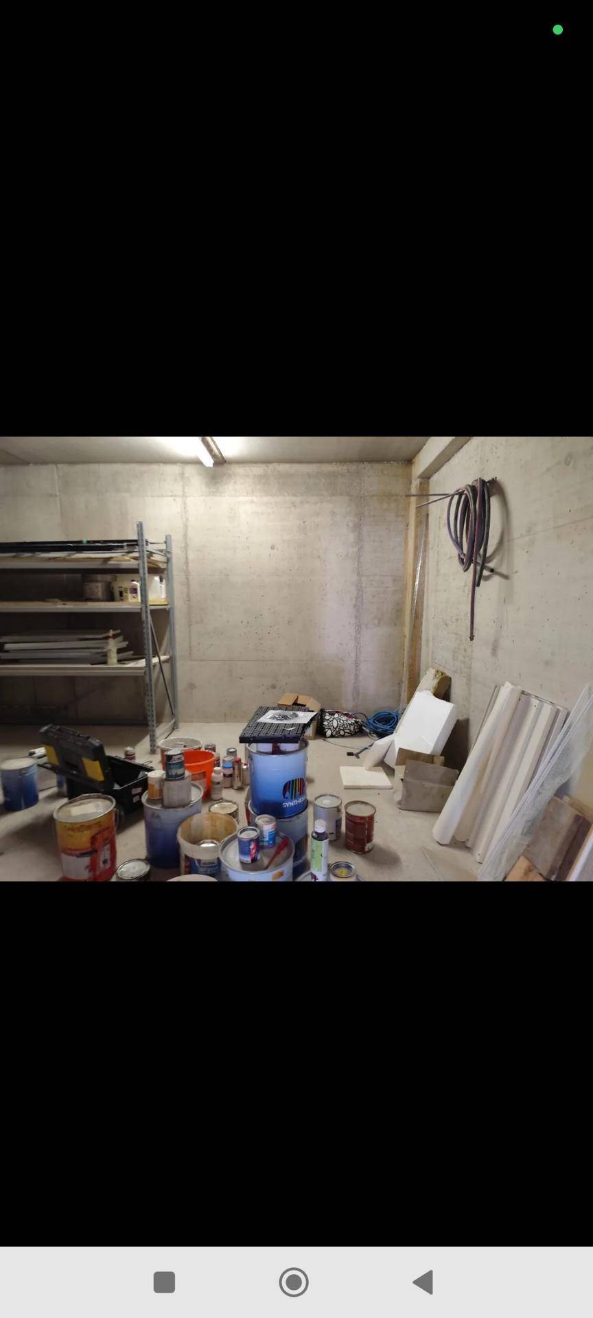 Kellerraum mit rohen Betonwänden, Metallregalen und zahlreichen Farbdosen auf dem Boden.