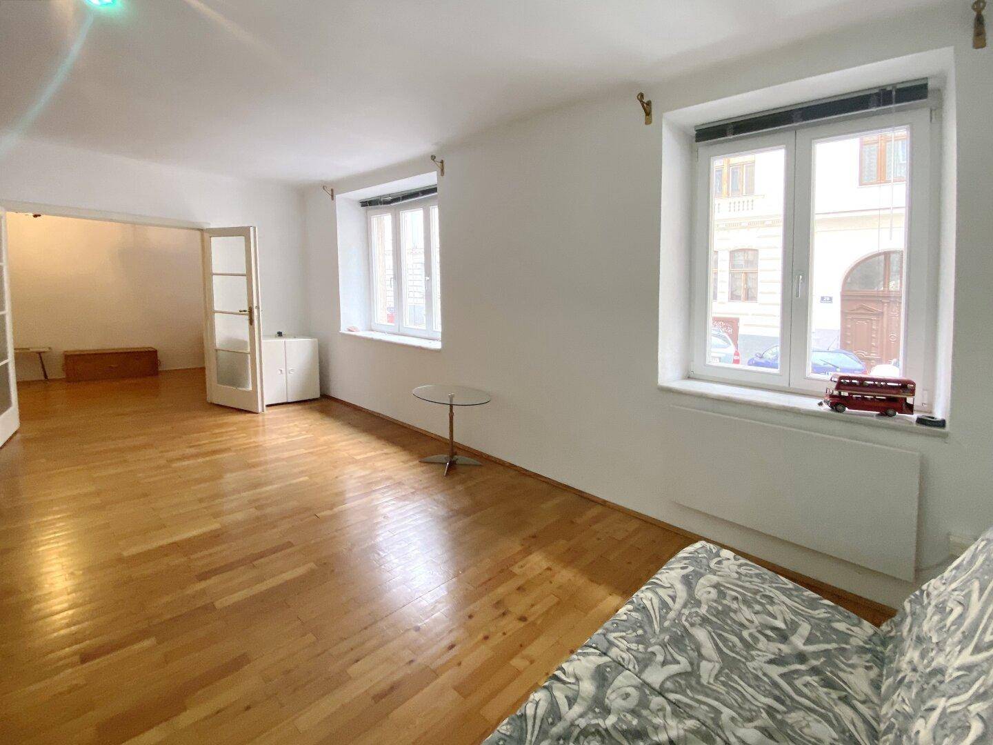 Helles Wohnzimmer mit Parkettboden und Blick auf die Straße, verbunden mit einem weiteren Raum.