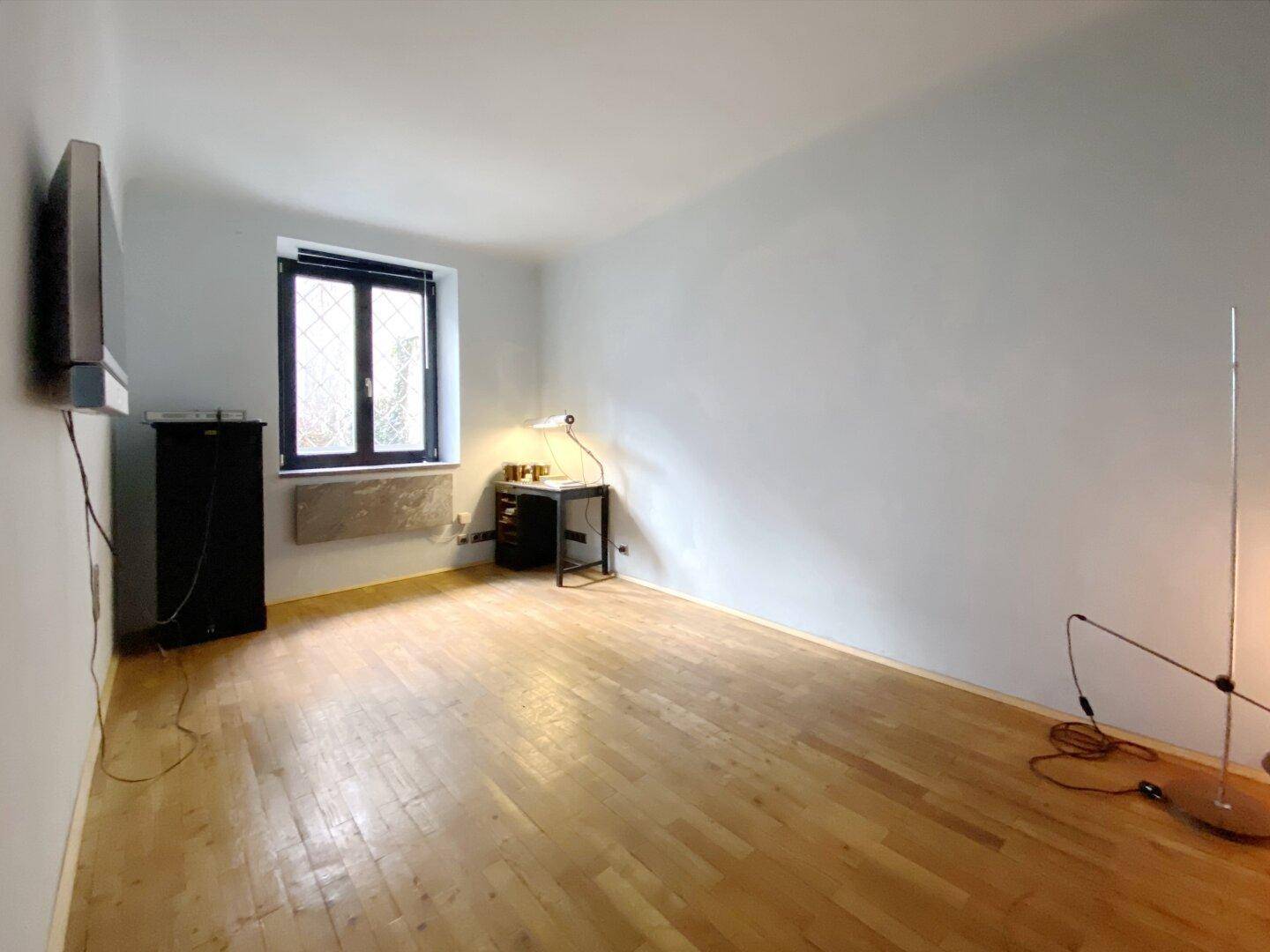 Heller Raum mit Parkettboden, einem Fenster und an der Wand montiertem Fernseher.