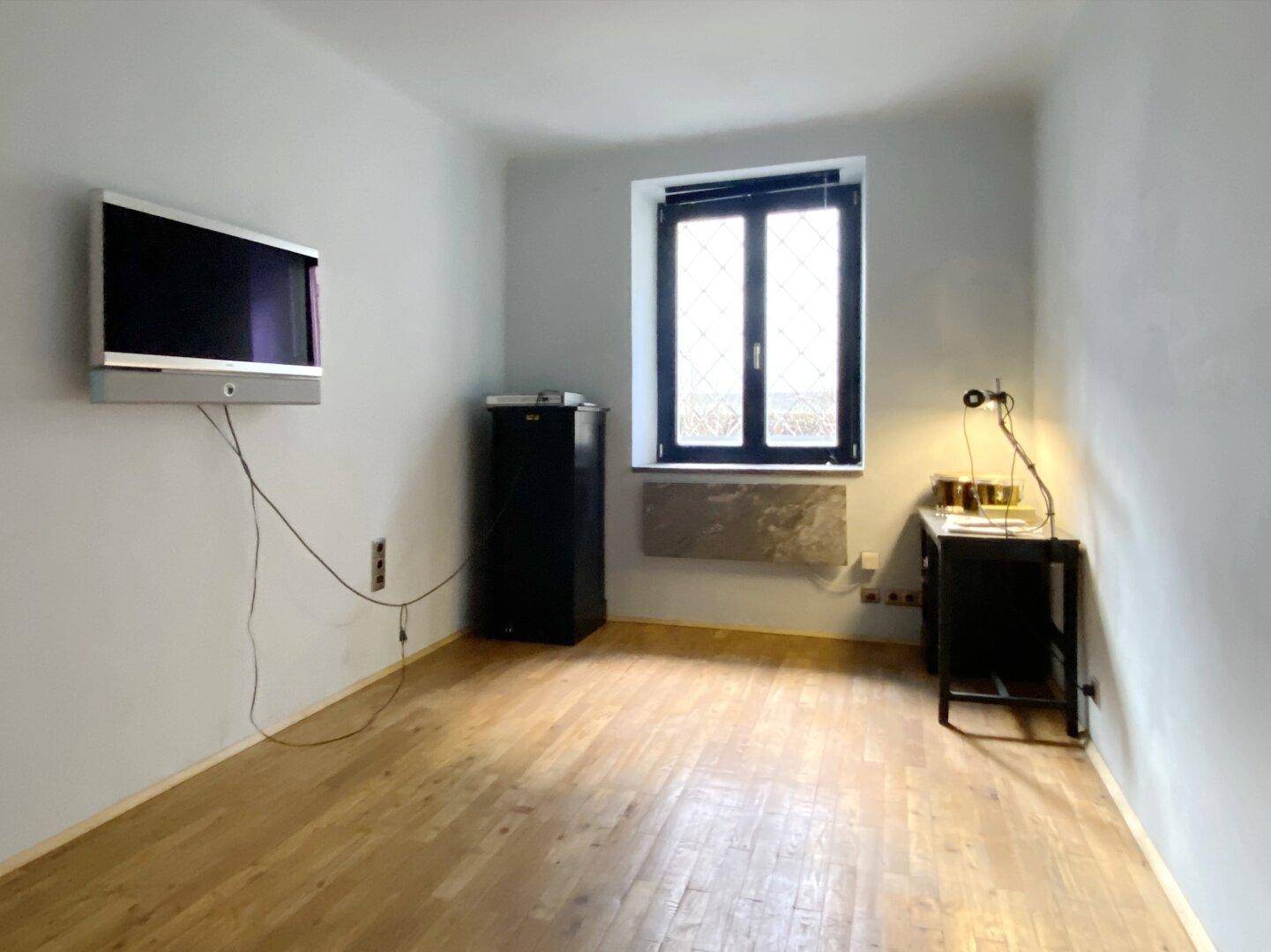 Schlichter Raum mit Holzboden, einem Fenster und einem an der Wand befestigten Flachbildfernseher.