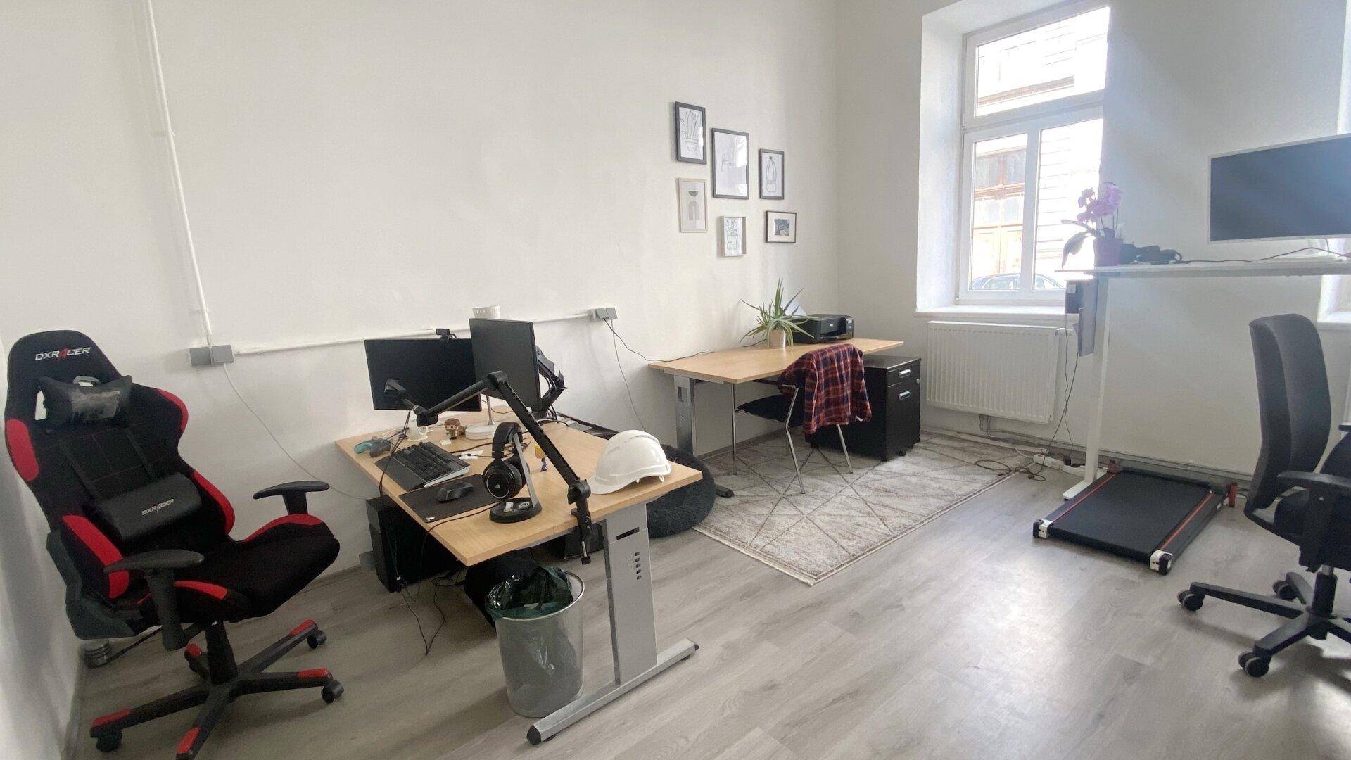 Heller Büroraum mit mehreren Schreibtischen, einem Gaming-Stuhl und Blick auf die umliegenden Gebäude.