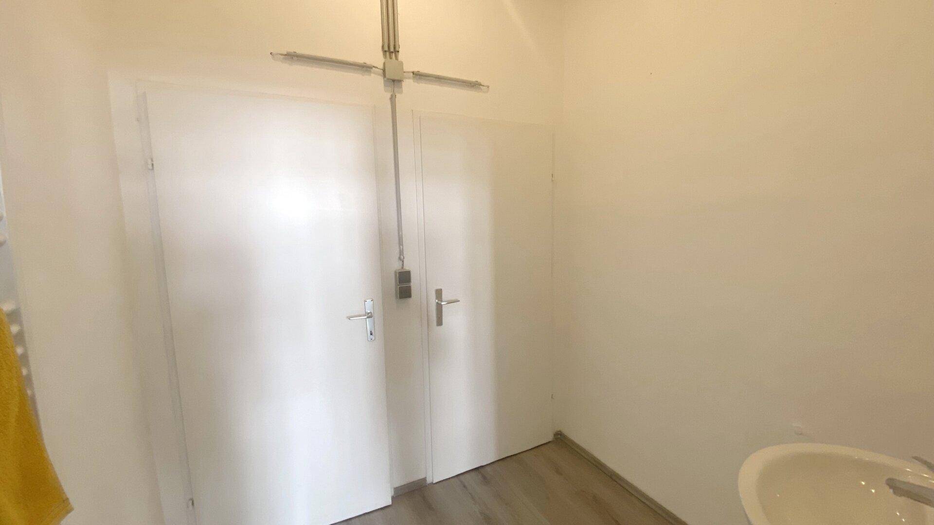 Zwei weiße Türen in einem schlichten Raum mit heller Beleuchtung und einem Waschbecken im Vordergrund.