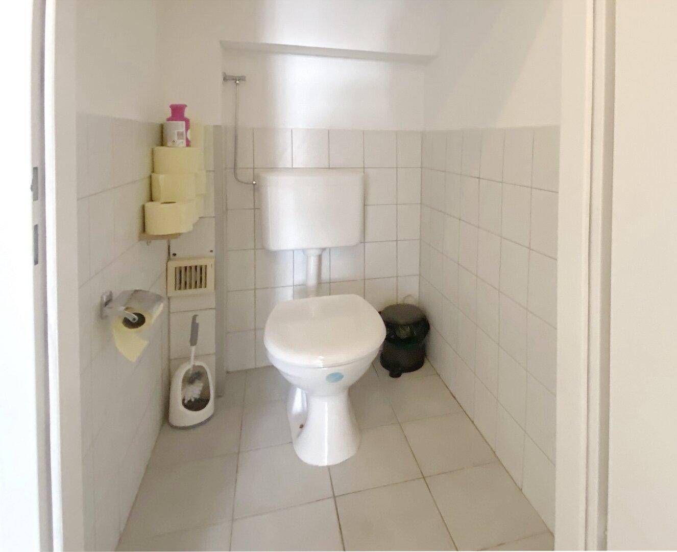 Einfache Toilette mit weißen Fliesen an den Wänden und auf dem Boden, sowie einem kleinen Mülleimer.