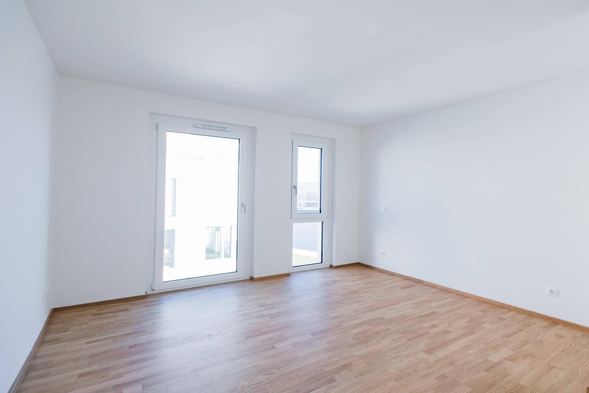 Helles Zimmer mit Holzboden und großen Fenstern, die viel Tageslicht hereinlassen.