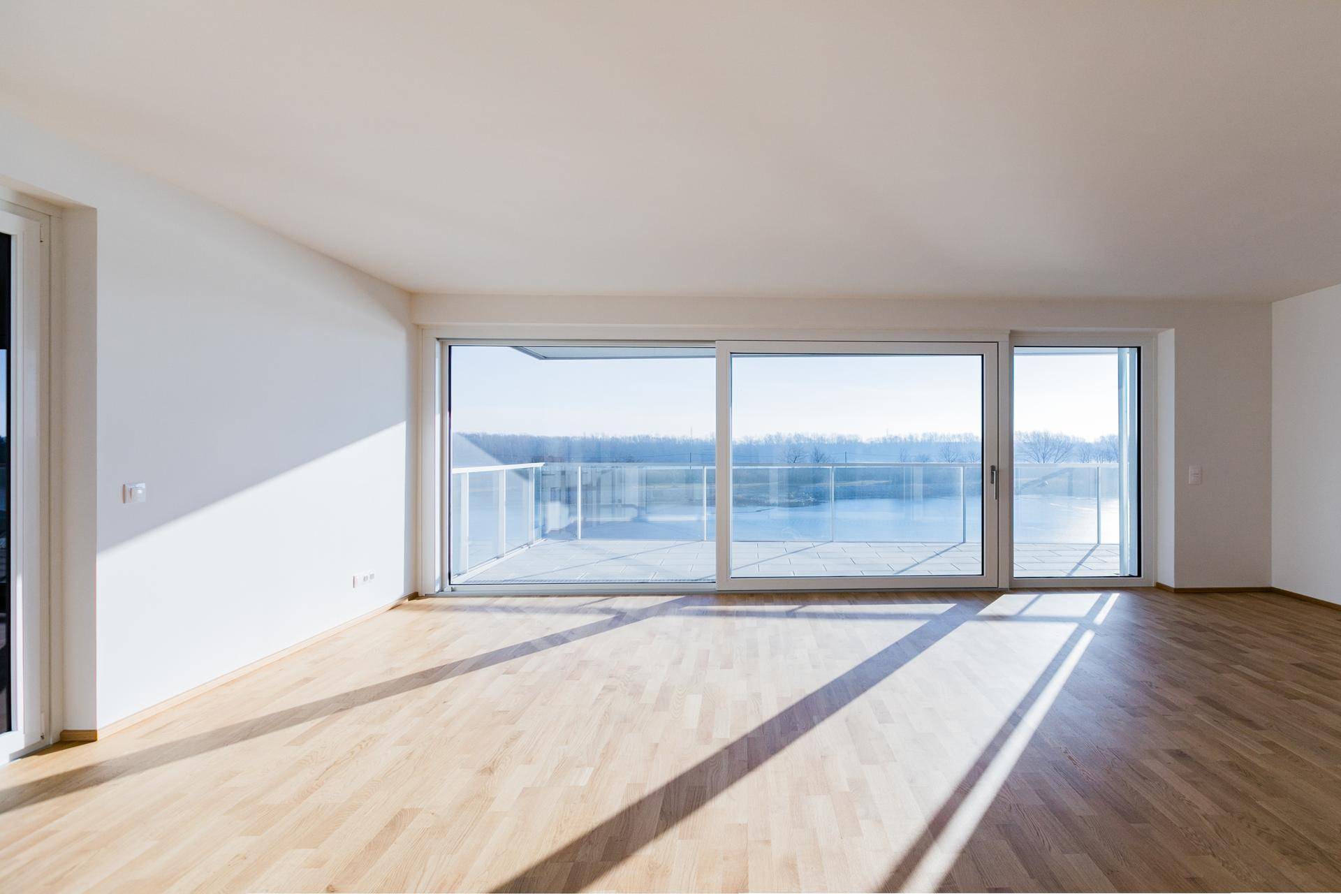 Geräumiges Wohnzimmer mit hellem Parkettboden und bodentiefen Fenstern mit Blick auf das Wasser.