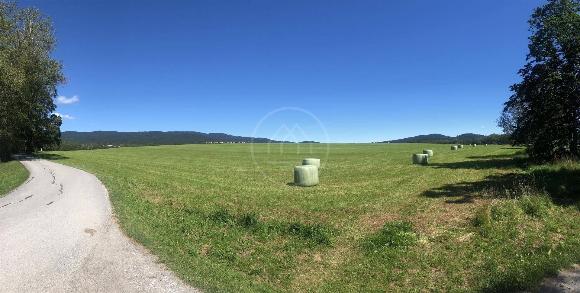 Weitläufige Bodenansicht eines grünen Feldes mit Heuballen, einer Straße und bewaldeten Hügeln am Horizont.
