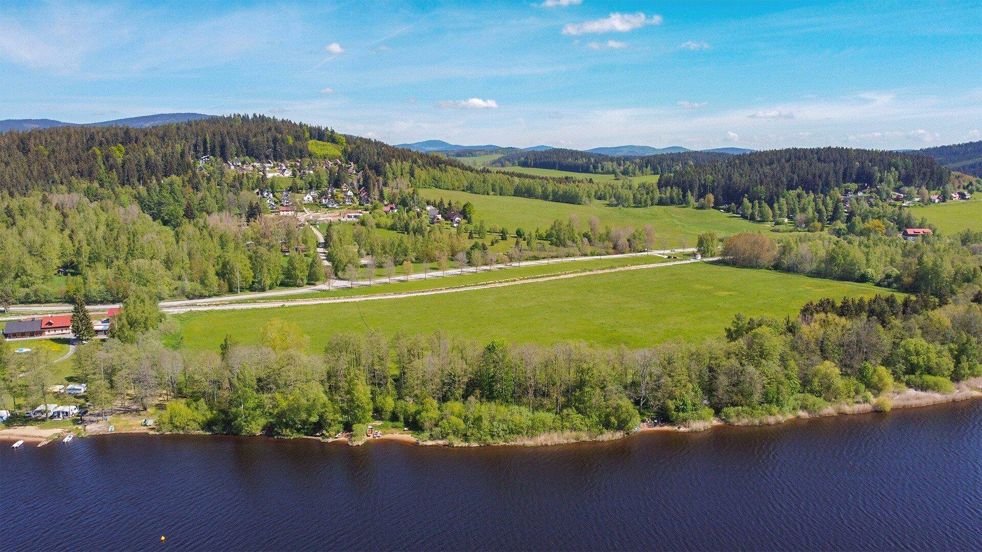 Weitläufige Landschaft mit einem See, grünen Wiesen und bewaldeten Hügeln unter blauem Himmel.