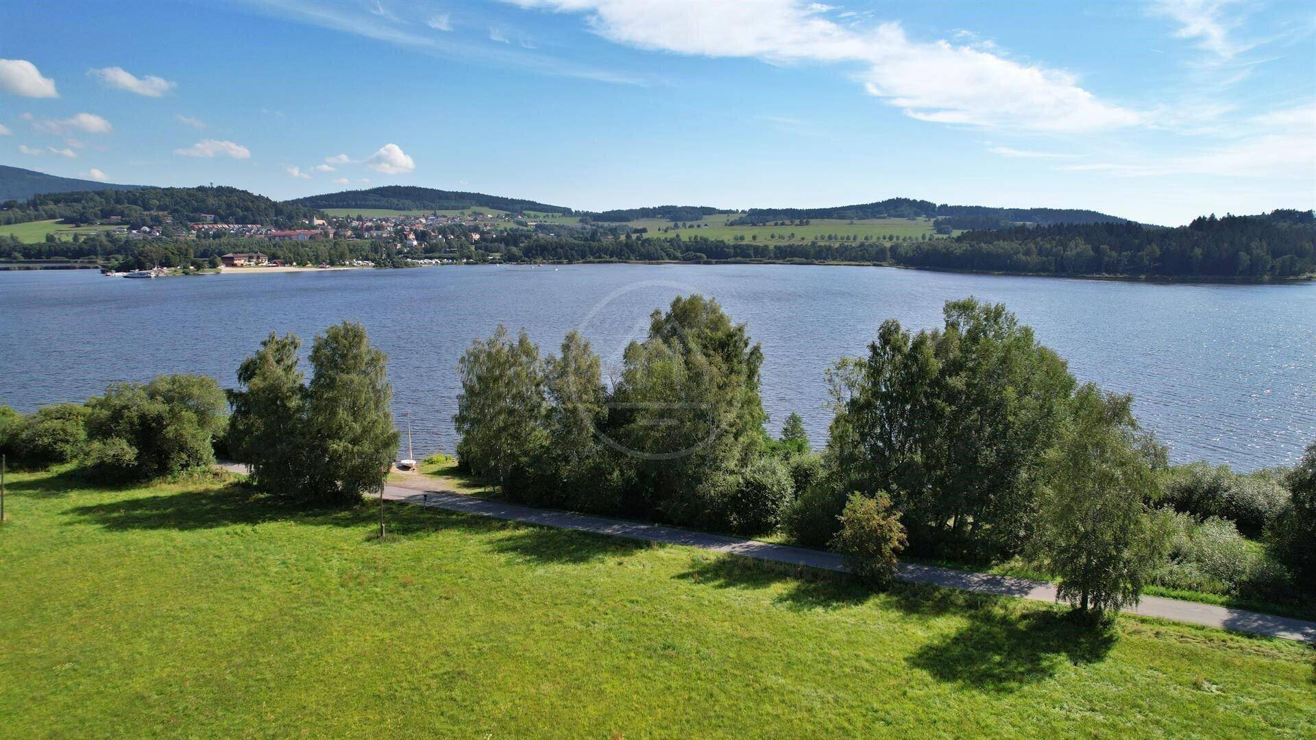 Blick auf den See mit einer Uferstraße, Bäumen und einem Dorf in der Ferne unter einem klaren blauen Himmel.