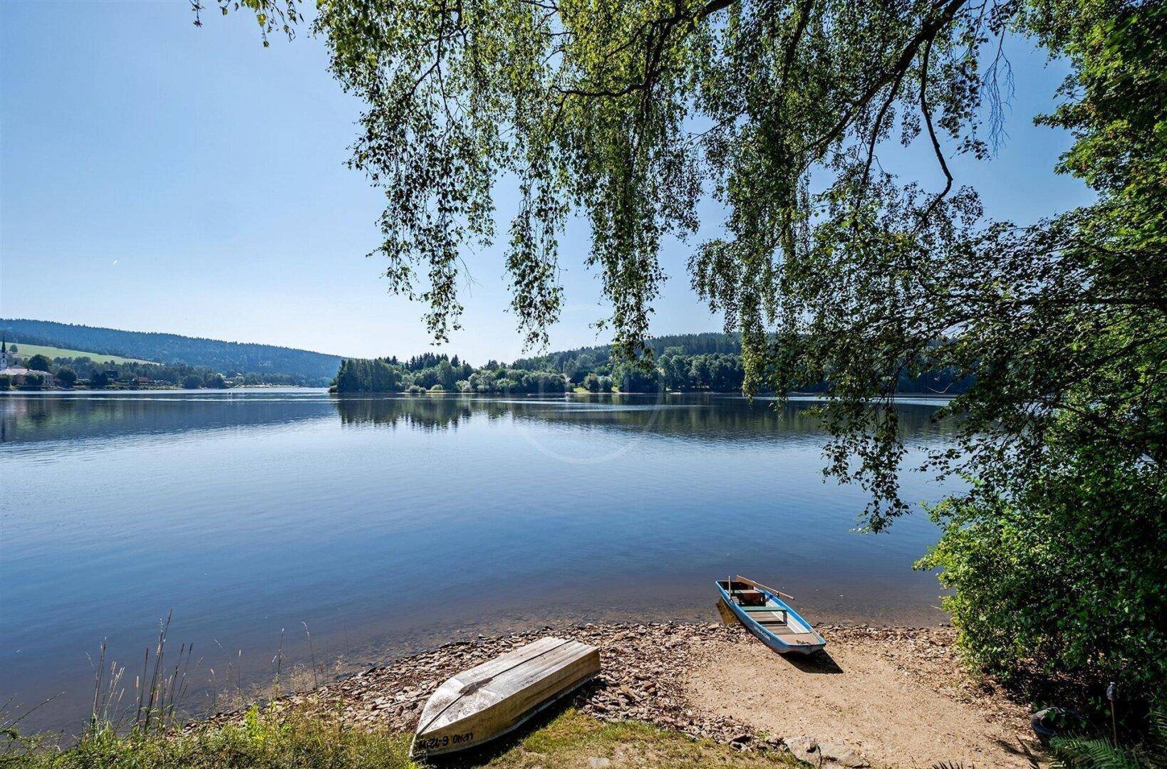 Idyllischer Seeblick mit zwei kleinen Booten am Ufer, umrahmt von grünen Bäumen und klarem Himmel.
