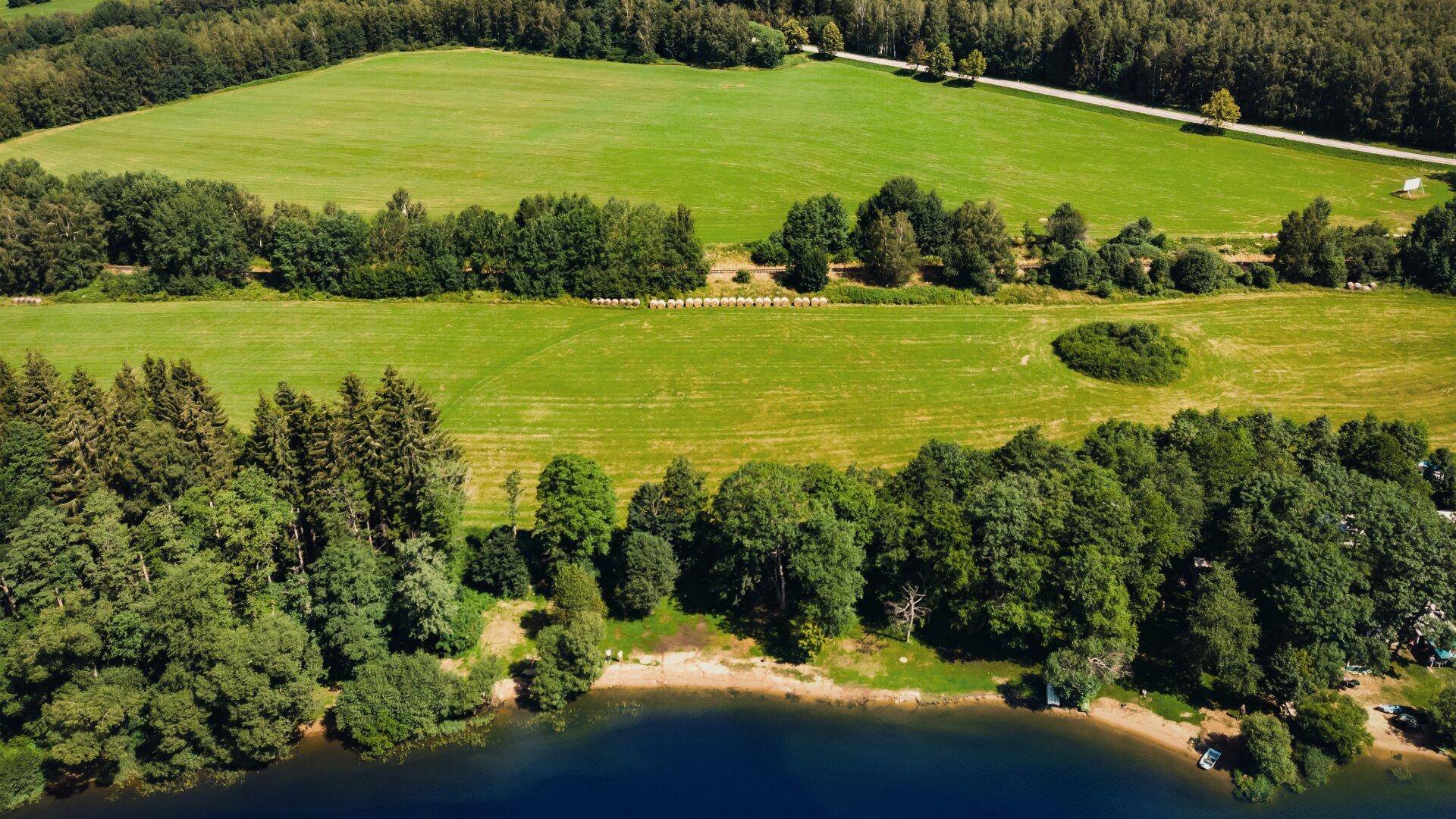 Luftaufnahme einer malerischen Landschaft mit See, Waldgebieten und weitläufigen grünen Feldern.