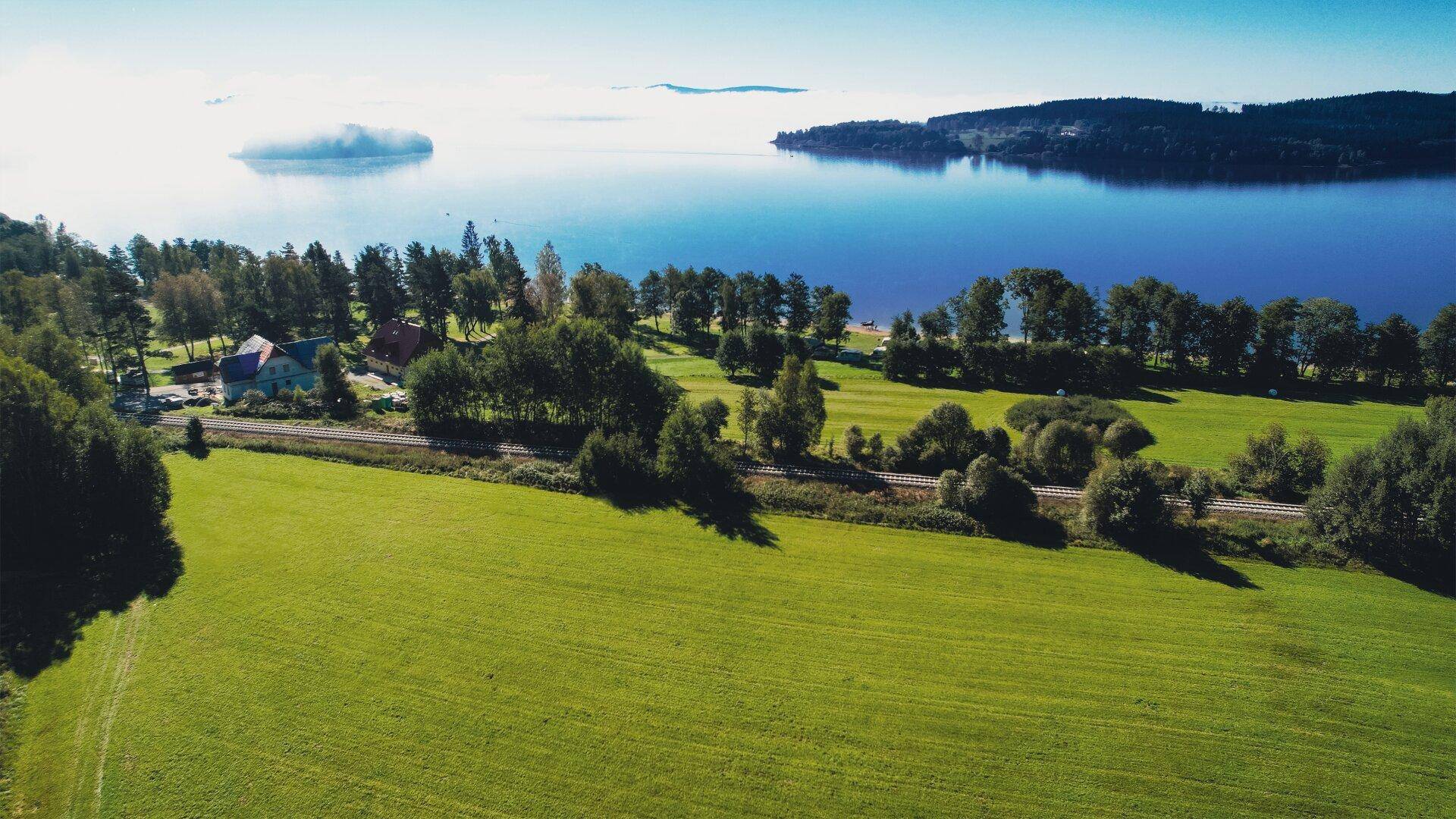 Luftaufnahme einer Landschaft mit See, grünen Feldern und einer Eisenbahnlinie am Horizont.
