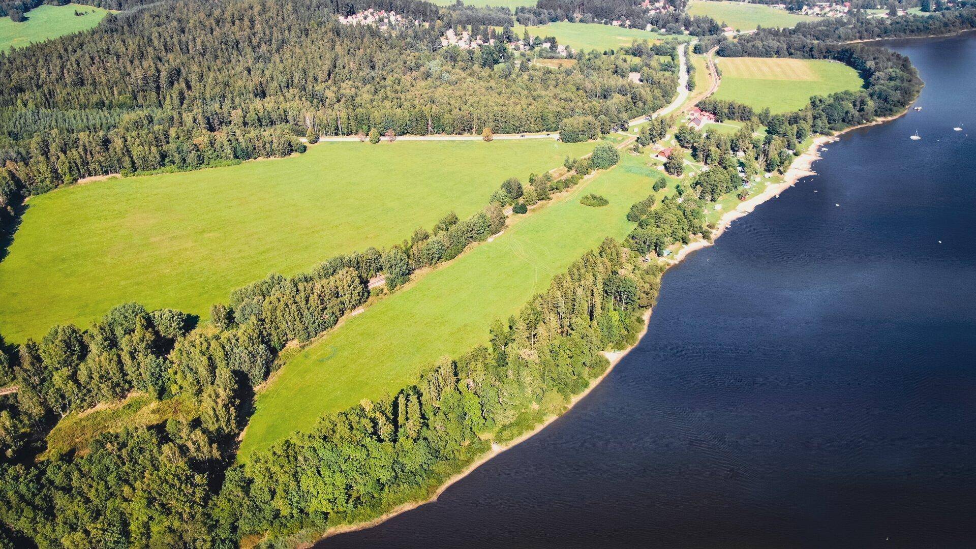 Panoramablick aus der Luft auf einen See, umgeben von dichten Wäldern und weiten Wiesen.