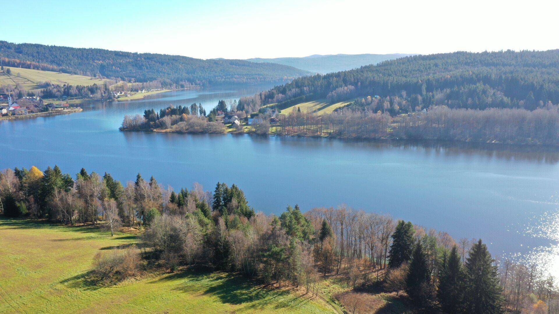Weitläufige Luftaufnahme eines Sees mit bewaldeten Ufern und einer kleinen Insel mit Häusern.