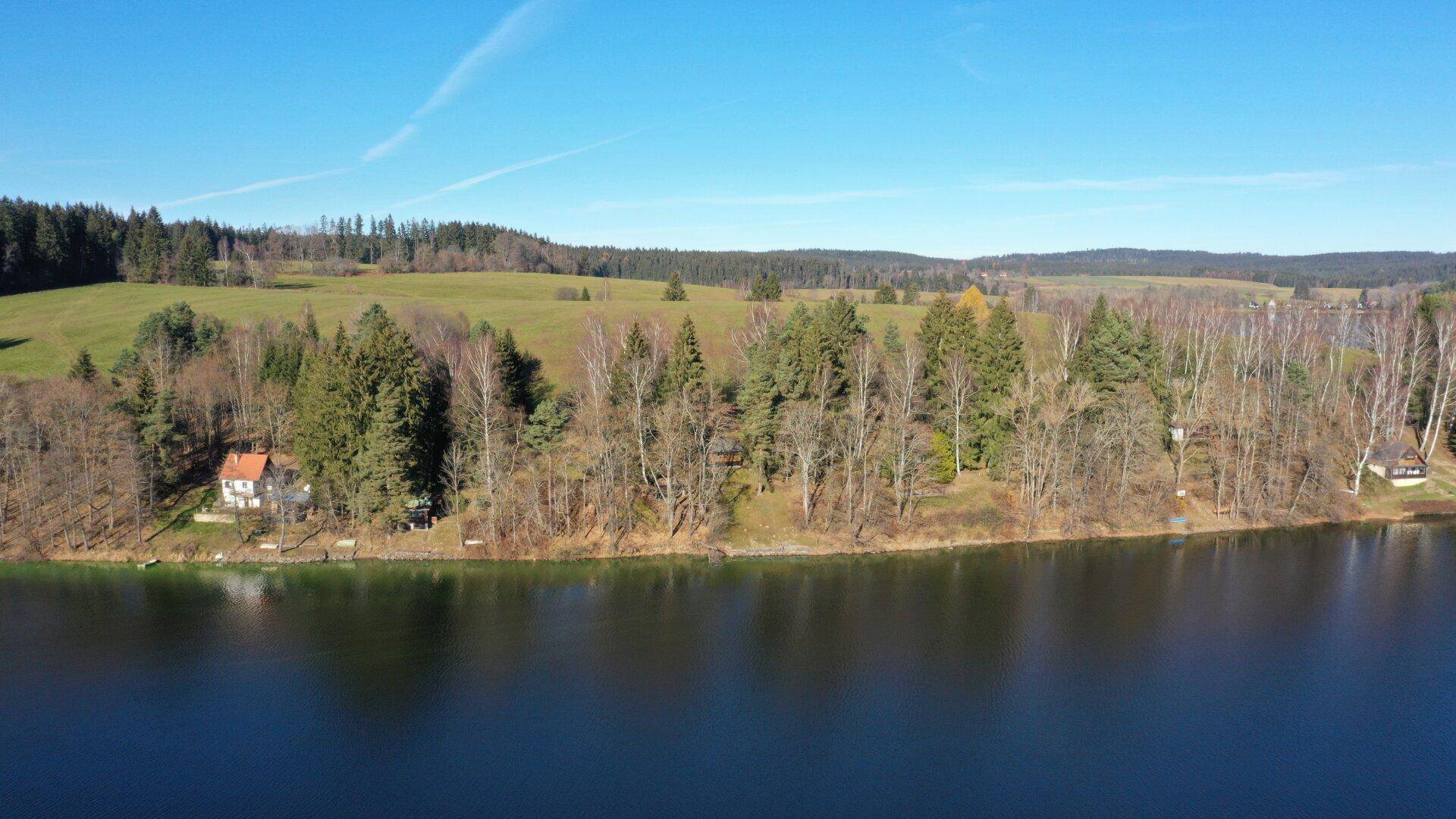 Luftaufnahme eines malerischen Sees umgeben von grünen Feldern und dichten Wäldern unter blauem Himmel.