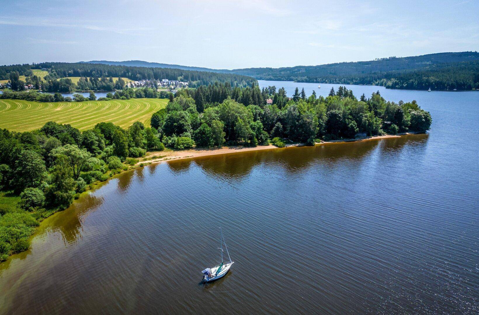 Luftaufnahme einer idyllischen Seenlandschaft mit bewaldeten Inseln und einem Segelboot.