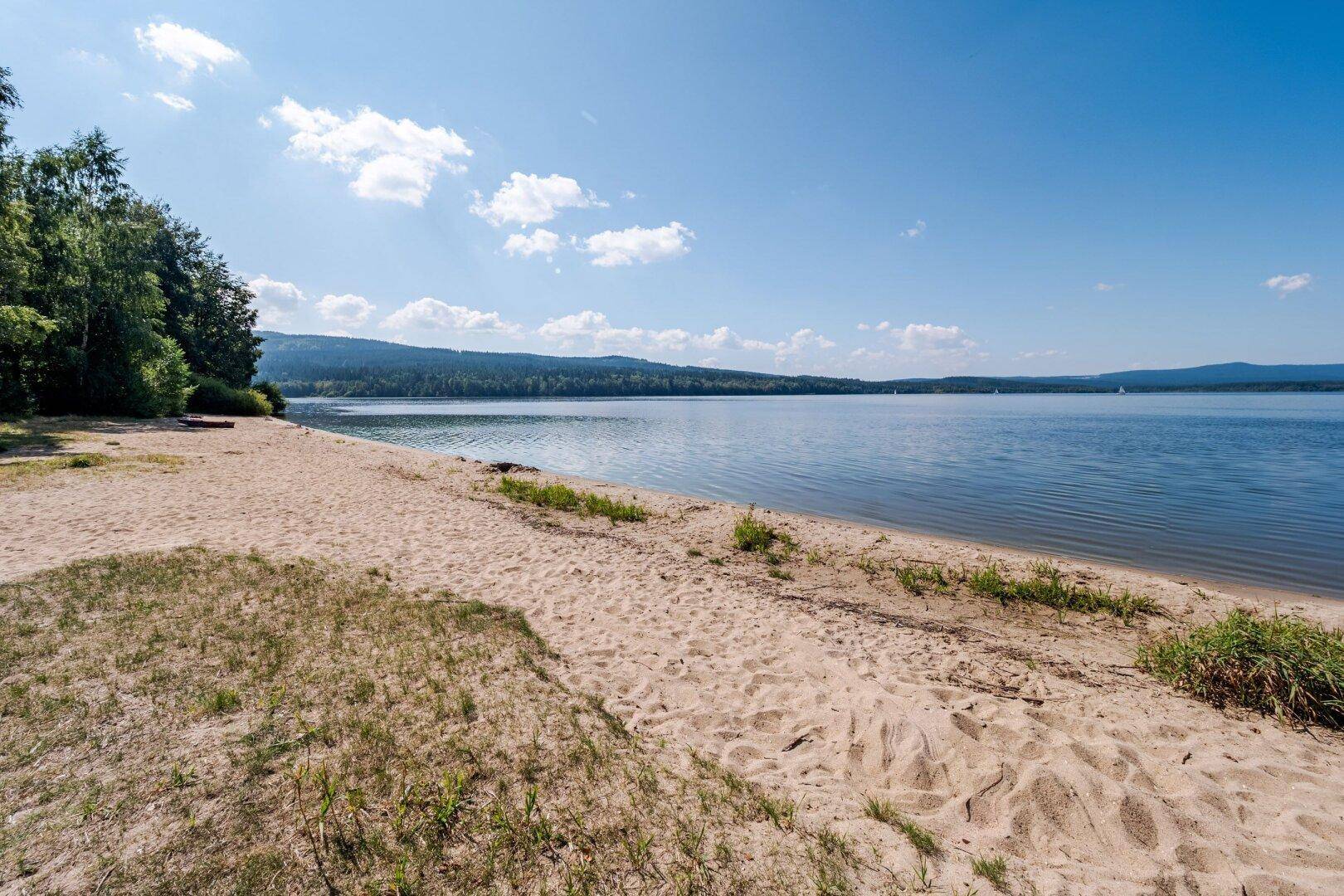 Sandstrand am Seeufer mit bewaldeten Hügeln im Hintergrund unter blauem Himmel.