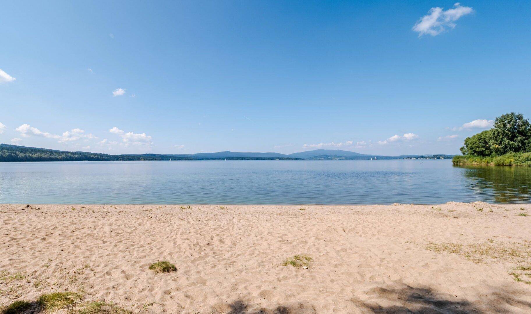 Weitläufiger Sandstrand am See mit klarem Wasser und bewaldeter Uferlinie.