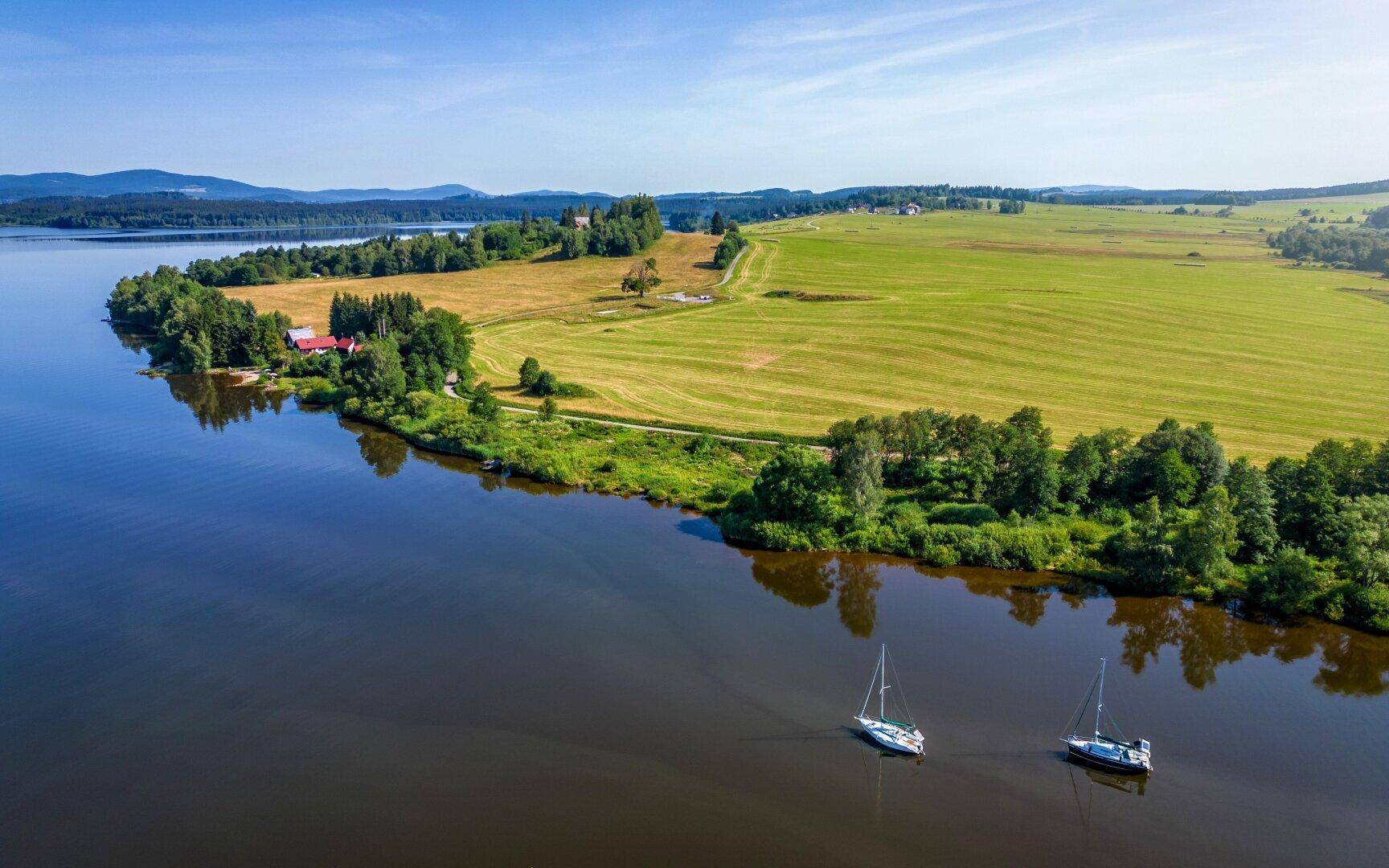 Weitläufige Landschaft mit einem See, grünen Feldern und bewaldeten Ufern unter blauem Himmel.