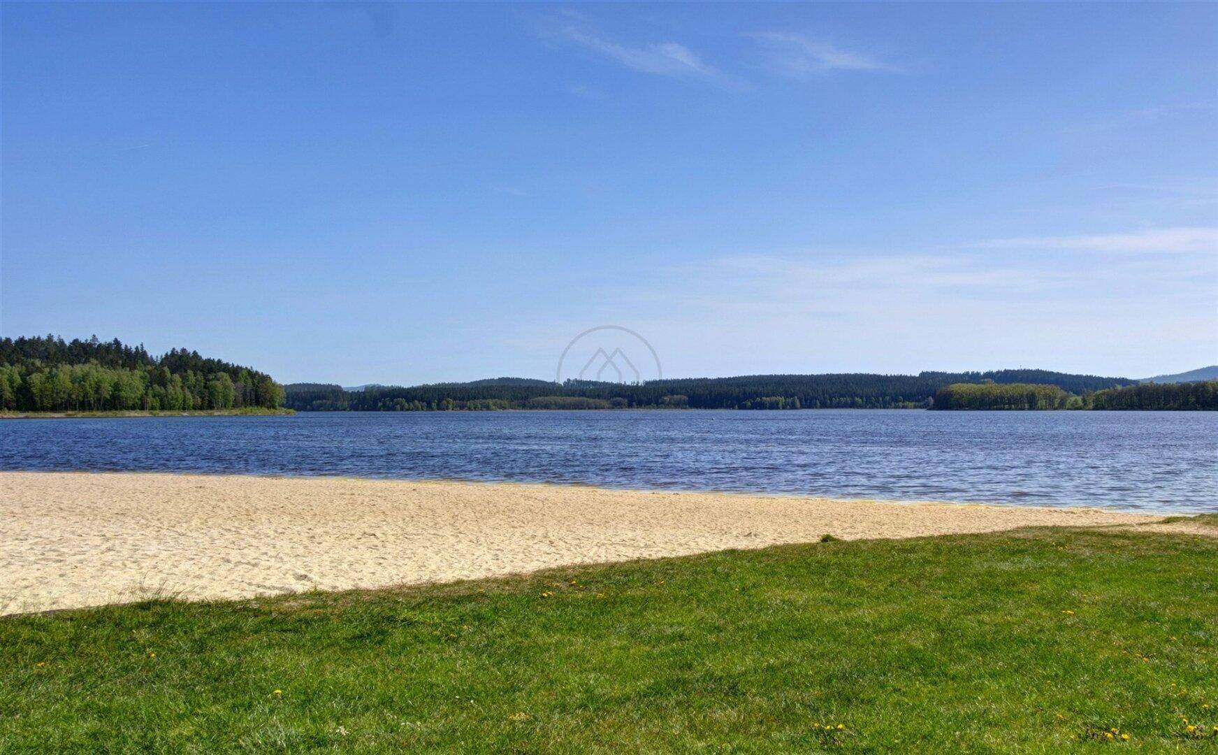 Breiter Sandstrand am Ufer eines großen Sees, umgeben von dichten Wäldern unter blauem Himmel.